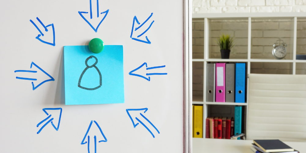 Sinnbild für Personalisierung: Post-it mit einem Männchen klebt mittig an einem Board in einem Büro, gemalte Pfeile zeigen von allen Seiten darauf.
