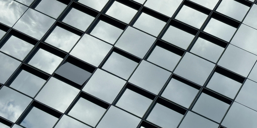 Raster aus quadratischen Glas‑ und Fassadenelementen eines modernen Gebäudes, die ein gleichmäßiges, modular aufgebautes Fenster‑ und Fassadenmuster bilden.